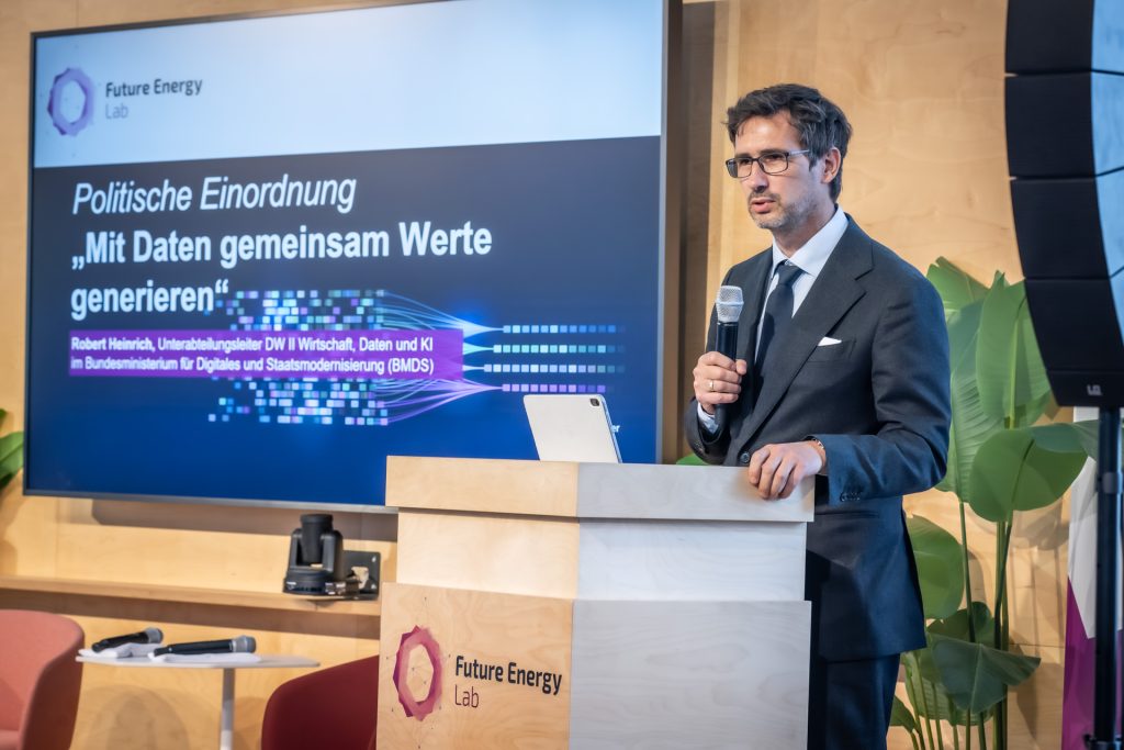 Mann mit Brille und Anzug hält Mikrofon und steht an einem Rednerpult mit Aufschrift 'Future Energy Lab' vor einem Bildschirm mit Text zu politischer Einordnung und Datenwerten