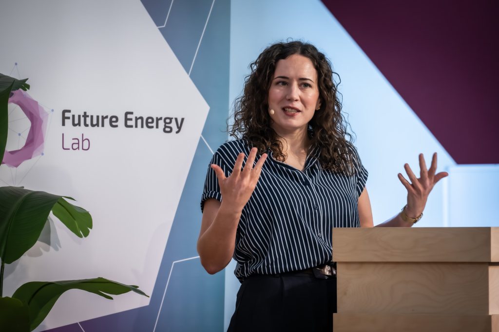 Frau mit lockigem Haar und gestreiftem Oberteil spricht gestikulierend an einem Rednerpult vor einem Banner mit der Aufschrift 'Future Energy Lab'
