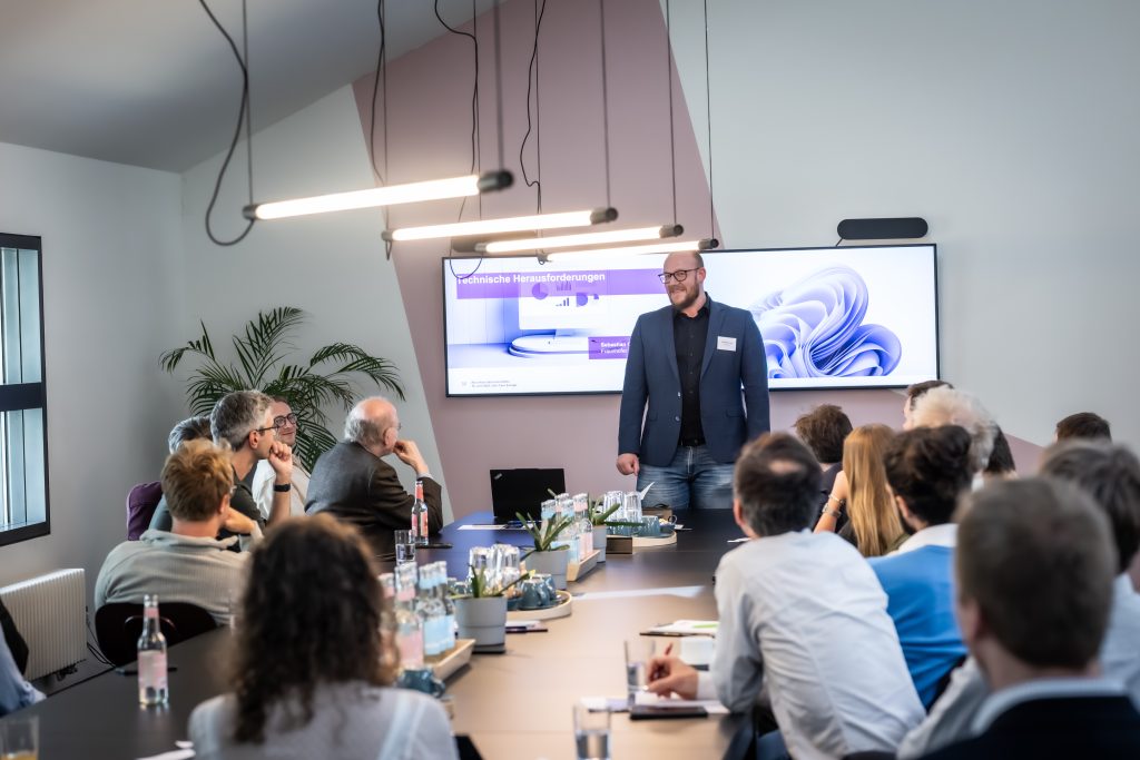 Mann steht vor Bildschirm mit Präsentation und spricht zu sitzendem Publikum in modernem Konferenzraum