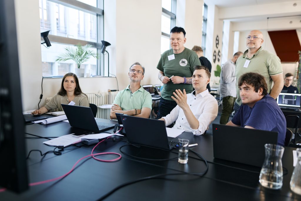 Fünf Personen sitzen und stehen an einem Tisch mit Laptops in einem hellen Büro und diskutieren konzentriert