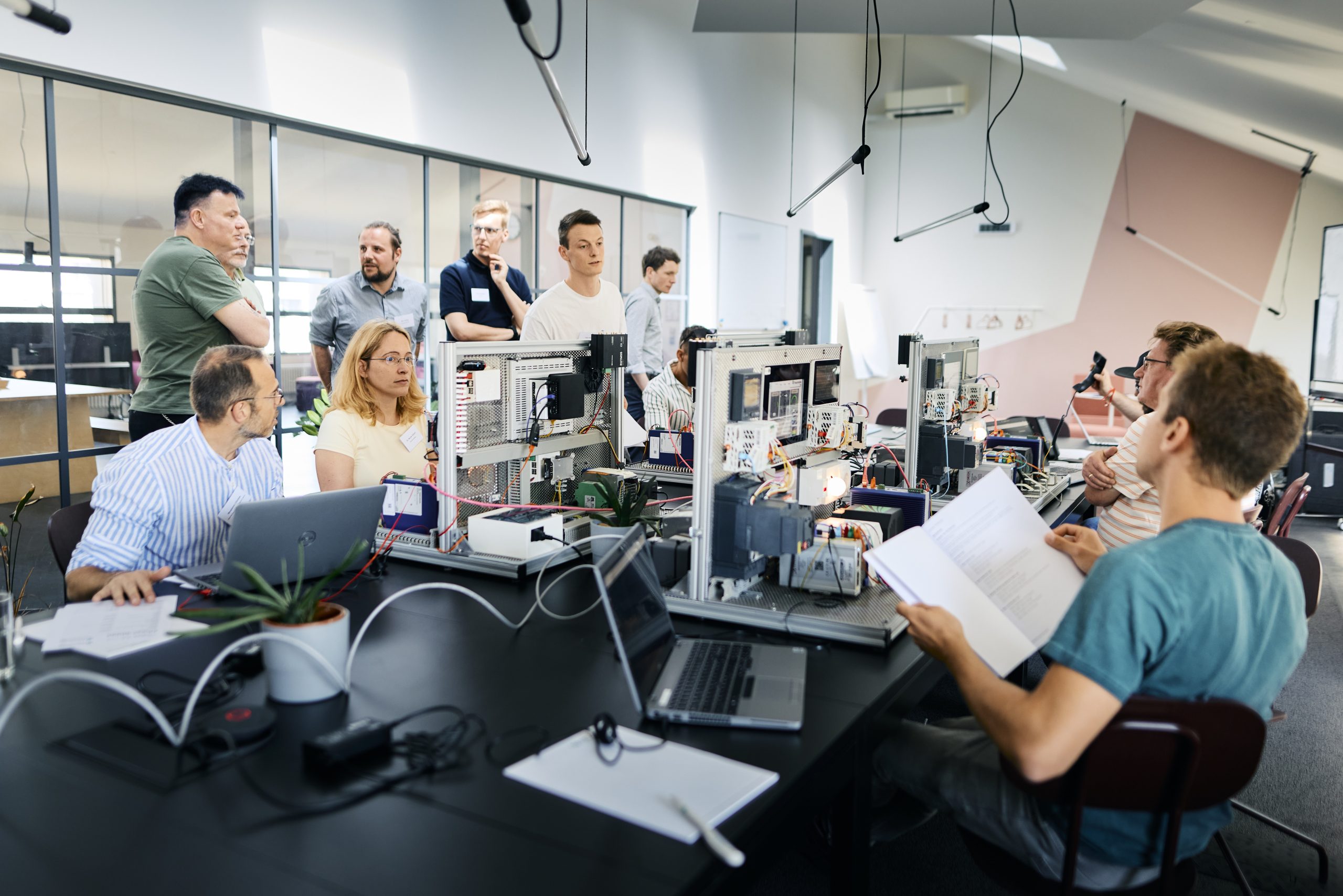 Mehrere Personen sitzen und stehen an einem langen Tisch mit Laptops und technischen Geräten in einem modernen Büro