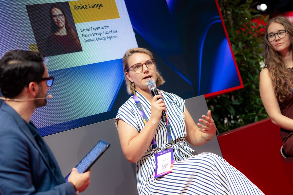 Frau mit Brille und gestreiftem Kleid spricht in Mikrofon während eines Podiumsgesprächs, Mann mit Brille und Mikrofon hält Tablet, weitere Frau sitzt daneben