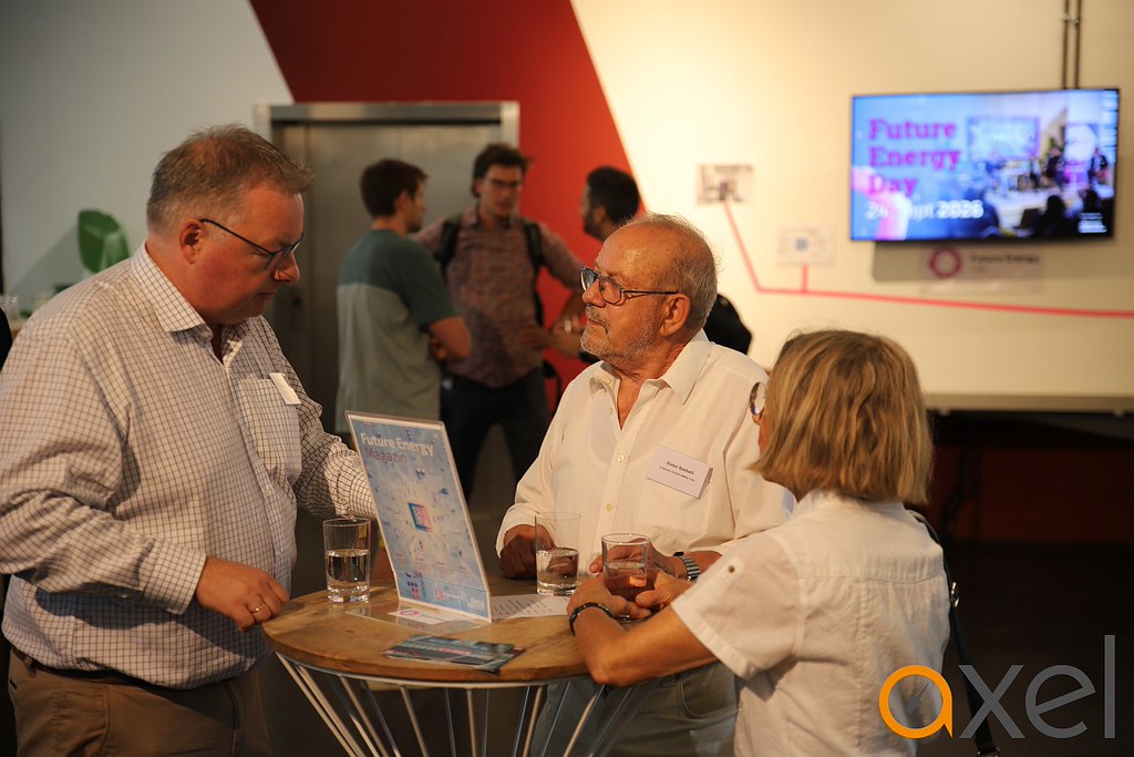 Drei Personen stehen an einem Stehtisch und unterhalten sich auf einer Veranstaltung mit einem Bildschirm im Hintergrund, auf dem 'Future Energy Day' zu lesen ist