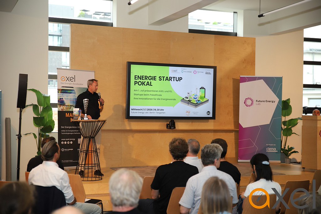 Mann mit Mikrofon steht vor Publikum und präsentiert auf großem Bildschirm eine Folie zum Energie Startup Pokal in modernem Veranstaltungsraum