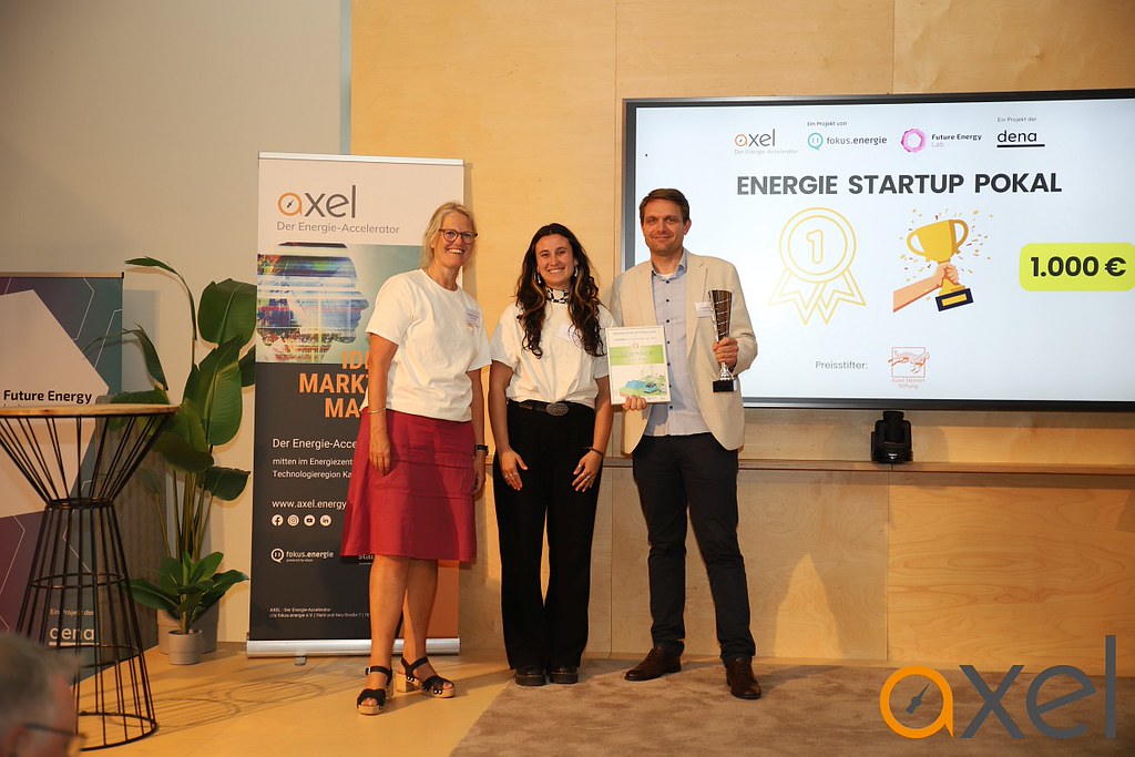 Drei Personen stehen vor einem Bildschirm mit der Aufschrift 'ENERGIE STARTUP POKAL' und einem Banner mit dem Logo 'axel', eine Person hält eine Urkunde und einen Pokal