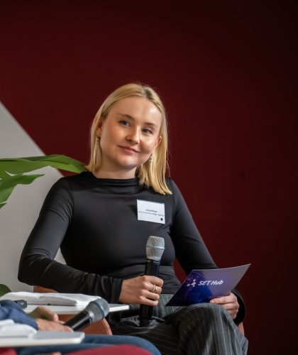 Frau mit blondem schulterlangem Haar in schwarzem Langarmshirt hält Mikrofon und SET Hub-Programmheft vor rotem Hintergrund