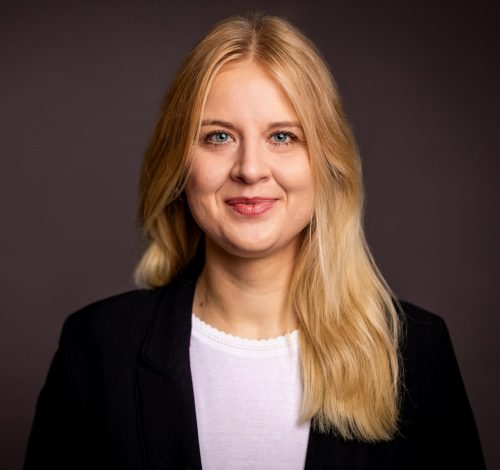 Frau mit langen blonden Haaren, weißem Oberteil und schwarzem Blazer vor dunklem Hintergrund lächelnd