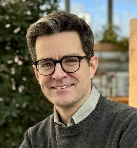 Porträt eines lächelnden Mannes mit dunkler Brille, grauem Hemd und dunklem Pullover vor unscharfem Hintergrund mit Pflanzen und Gebäuden