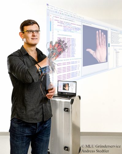 Mann mit Brille trägt schwarze Hemd und hat Sensorhandschuh mit Kabeln an der rechten Hand vor einem Laptop und einer Projektion von Hand und Programmcode