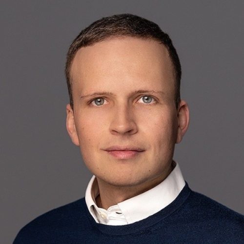 Porträt eines Mannes mit kurzem Haar, weißem Hemd und dunkelblauem Pullover vor grauem Hintergrund