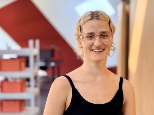 Frau mit blonden, schulterlangen Haaren, die eine schwarze ärmellose Bluse und eine Brille trägt, lächelt in einem hellen Raum mit Regalen im Hintergrund